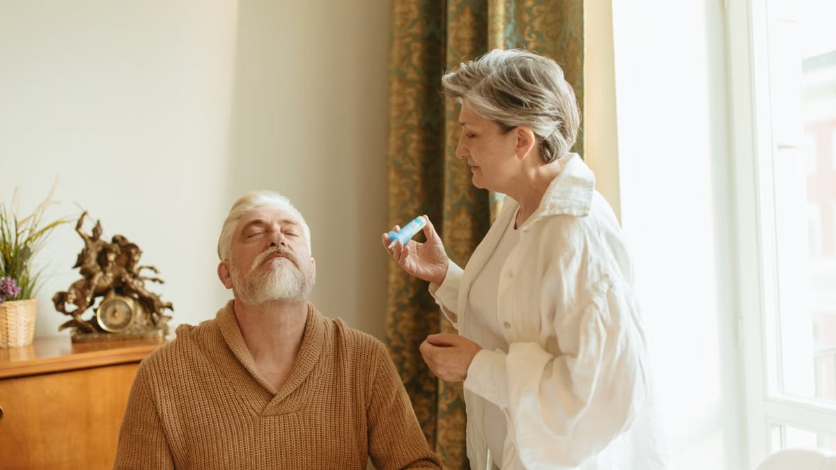 An elderly woman assists her husband with health care indoors by the window.